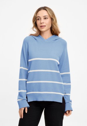 Frau mit hellbraunen Haaren trägt einen hellblauen Kapuzenpullover mit weißen horizontalen Streifen und schwarze Hose, sie steht vor einem weißen Hintergrund.