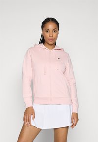 Haut zipé rose en tissu doux, doté d'une capuche, de cordons de serrage, de poches kangourou et d'un petit logo sur la poitrine gauche.