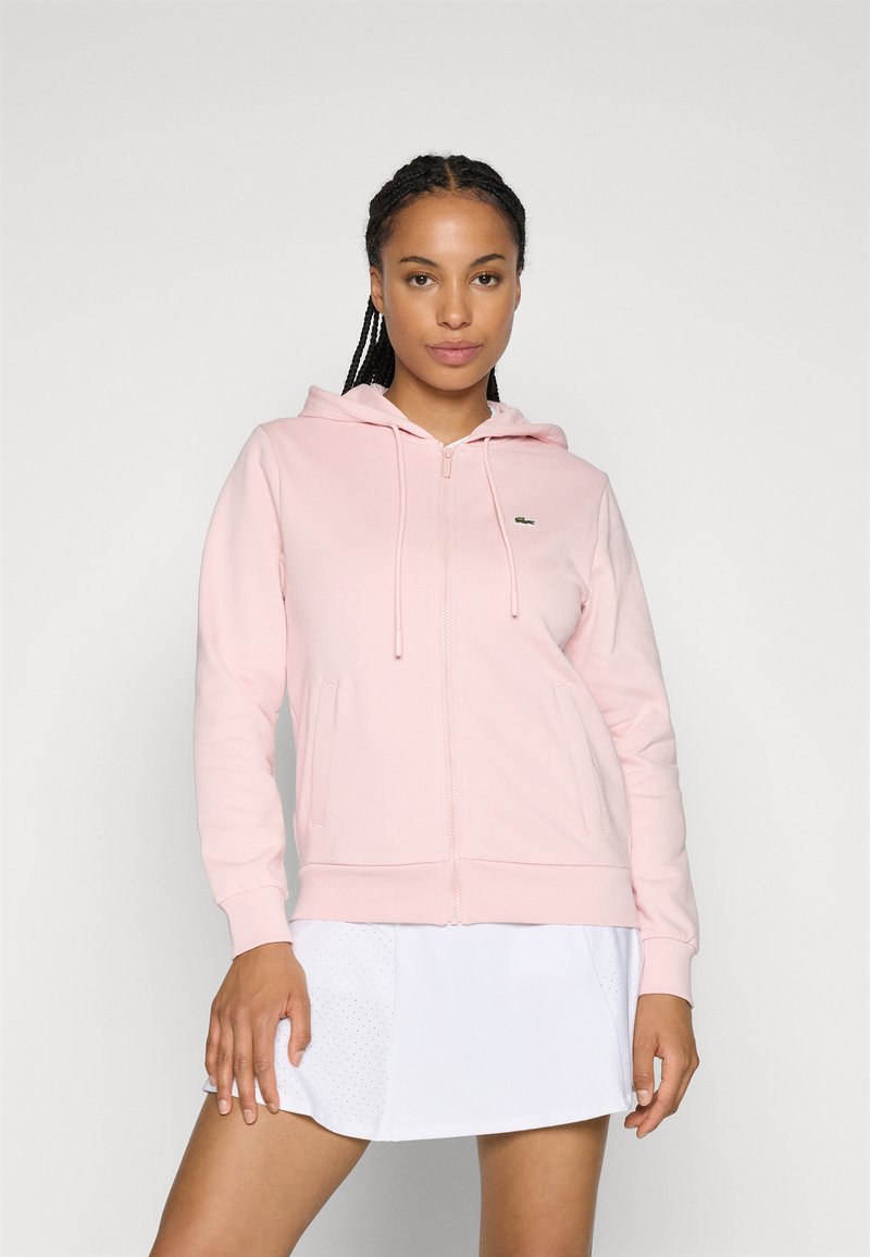Haut zipé rose en tissu doux, doté d'une capuche, de cordons de serrage, de poches kangourou et d'un petit logo sur la poitrine gauche.