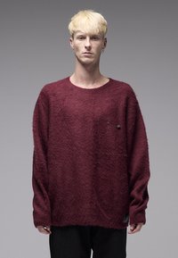 Μπορντό, φαρδύ πουλόβερ με αφράτη υφή, φαρδιά λαιμόκοψη και διακοσμητική παραμάνα μπροστά; συνδυασμένο με μαύρο παντελόνι.