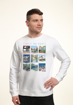 Młody mężczyzna w białej bluzie z kolorowym napisem „Emily in Paris” i dziewięcioma ilustrowanymi scenami z Paryża w siatce 3x3, patrzy w bok.