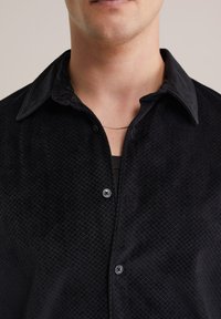 Camicia nera con bottoni, caratterizzata da un motivo testurizzato, con colletto classico e bottoni blu. Il tessuto appare morbido e leggermente lucido.