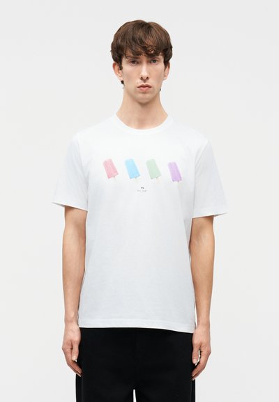 Νεαρός άνδρας που φοράει λευκό T-shirt με τέσσερις παστέλ αποχρώσεις παγωτού ξυλάκι και μαύρο παντελόνι, στέκεται μπροστά σε ένα απλό φόντο.