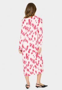 Rochie lungă, plisată, în nuanțe de roz și alb cu un model tie-dye. Prezintă mâneci lungi și o lungime medie. Este asortată cu balerini negri.