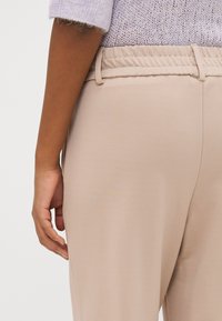 Beige stretchy pants with a smooth texture, elastic waistband, and side seam details, shown alongside a partially visible hand.