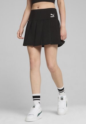 Kobieta nosząca czarną plisowaną spódnicę Puma, białe sneakersy Puma oraz białe skarpetki z czarnymi paskami, stoi na tle jednolitego szarego tła.
