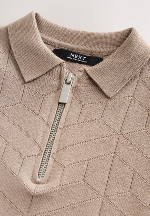 Gaiši brūns polo krekls ar rāvējslēdzēju, ar teksturētu ģeometrisku rakstu, īsu apkakli un sudraba krāsas rāvējslēdzēju. Zīmola birka redzama apkakles iekšpusē.