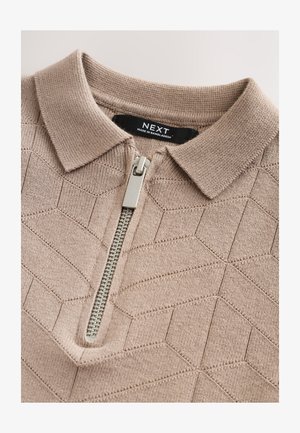 Jasnobrązowa koszulka polo zapinana na zamek z teksturowanym geometrycznym wzorem, krótkim kołnierzykiem i srebrnym suwakiem. Wewnątrz kołnierzyka widoczna metka marki.