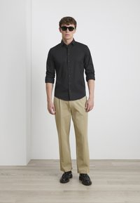 Schwarze Knopfhemd, hochgekrempelte Ärmel, kombiniert mit beigen Hosen und schwarzen Schuhen. Mann mit dunkler Sonnenbrille steht vor einer schlichten Wand.
