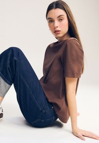 Vrouw met lang bruin haar zit op de vloer, draagt een bruin shirt met korte mouwen, omgeslagen donkere spijkerbroek, grijze sokken en witte sneakers tegen een effen achtergrond.