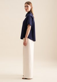 Granatowa bluzka z krótkim rękawem wykonana z gładkiego materiału, zestawiona z wysokimi, prostymi spodniami w kolorze kremowym i czarnymi sandałami na odsłoniętych palcach.
