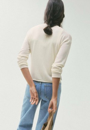 Pull couleur crème avec texture côtelée, associé à un jean bleu clair. Le pull a des manches longues et un design simple et classique.