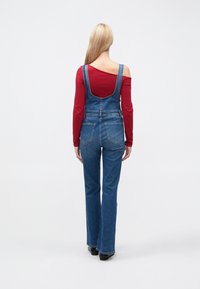 Salopette in denim blu slavato, con bretelle regolabili, gambe svasate e tasche posteriori, indossata sopra una maglia rossa a maniche lunghe.