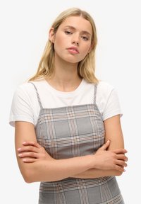 Junge Frau mit blonden Haaren, die die Arme verschränkt, trägt ein weißes Kurzarmoberteil unter einem karierten ärmellosen Kleid, neutrale Mimik, schlichter Hintergrund.