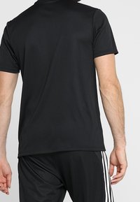 Černé sportovní tričko z hladké látky, s kulatým výstřihem a krátkými rukávy. Nošeno s odpovídajícími černými šortkami s bílými pruhy.