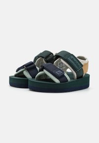 Sandale bicolore cu curele de culoare verde închis și navy, talpă texturată de culoare verde și o talpă ușoară cu un fundal albastru în contrast.