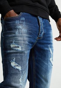 Jeans de mezclilla en un color azul desgastado que presentan parches desgastados y bordes deshilachados. La tela muestra variaciones en textura y tono.