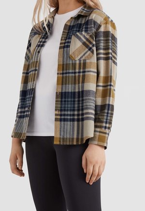 Kvinde iført en åben ternet flannelskjorte i beige, marineblå og sennepsfarvet over en hvid skjorte med sorte bukser mod en ensfarvet baggrund.