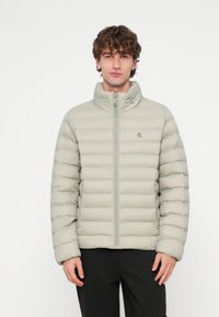 Casaco puff cinza claro com design acolchoado horizontal, gola alta, fecho de correr frontal e detalhe do logótipo no peito. O tecido parece liso e suave.