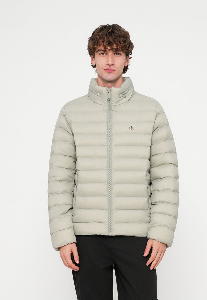 Casaco puff cinza claro com design acolchoado horizontal, gola alta, fecho de correr frontal e detalhe do logótipo no peito. O tecido parece liso e suave.