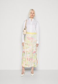 Weißes Hemd mit Knöpfen, kombiniert mit einem plissierten, blumigen Maxirock in Pastellfarben und einem schmalen gelben Bund, ergänzt durch weiße Absätze und eine beige Tasche.