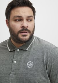 Grå polo skjorte med teksturert mønster, med knappet krage og hvit brodert logo på brystet. Har kontrasterende kragebeskytter.