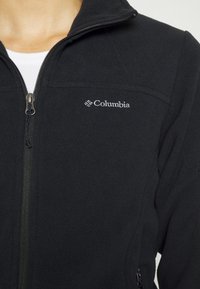 Mustat fleece-takki, jossa on vetoketjullinen etuosa ja korkea kaulus. Columbia-logo on brodeerattu vasempaan rintaan. Pehmeä pinta.