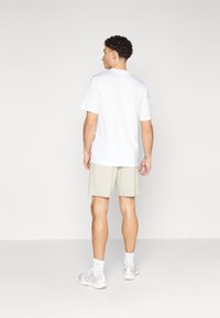 T-shirt blanche à manches courtes portée avec un short beige clair. Les chaussures du modèle sont grises claires avec des accents blancs. Design simple et décontracté.