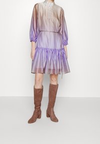 Robe dégradé violet avec des manches ballons, un col boutonné et une taille froncée. Associée à des bottes en daim marron jusqu'au genou.