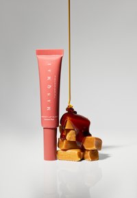 Rosa läppglans i tub bredvid karamellgodis, med glansig karamellsoja som rinner ner och framhäver släta yttexter och varma toner.