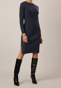 Abito in maglia a coste navy con maniche lunghe e spacchi laterali, abbinato a stivali in pelle nera al ginocchio. Texture liscia e design aderente.