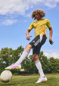 Sportoló sárga és piros Adidas mezben, fekete sortban, fehér zokniban és rózsaszín részletekkel ellátott stoplis cipőben, foci rúgást hajt végre egy füves pályán.