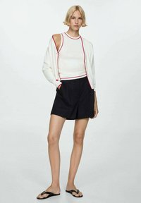 Donna bionda che indossa un top senza maniche bianco con rifiniture rosse, un cardigan bianco, pantaloni corti neri e ciabatte nere, in piedi contro uno sfondo bianco.
