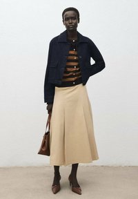 Marineblaue Jeansjacke über einem braun-schwarz gestreiften Pullover, kombiniert mit einem beigen Faltenrock und braunen High Heels, während sie eine braune Tasche hält.