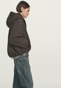 Persona con i capelli corti che indossa un giubbotto imbottito scuro con cappuccio, con le mani nelle tasche e jeans blu larghi, in piedi di profilo.