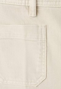 Gros plan d'une poche arrière en jean beige avec des coutures visibles et une passants de ceinture au-dessus sur un tissu texturé.