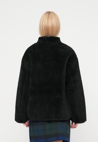 Chaqueta de forro polar negra con cuello alto, mangas anchas y acabado texturizado. Dobladillo inferior de cuadros visible de una prenda superpuesta debajo.