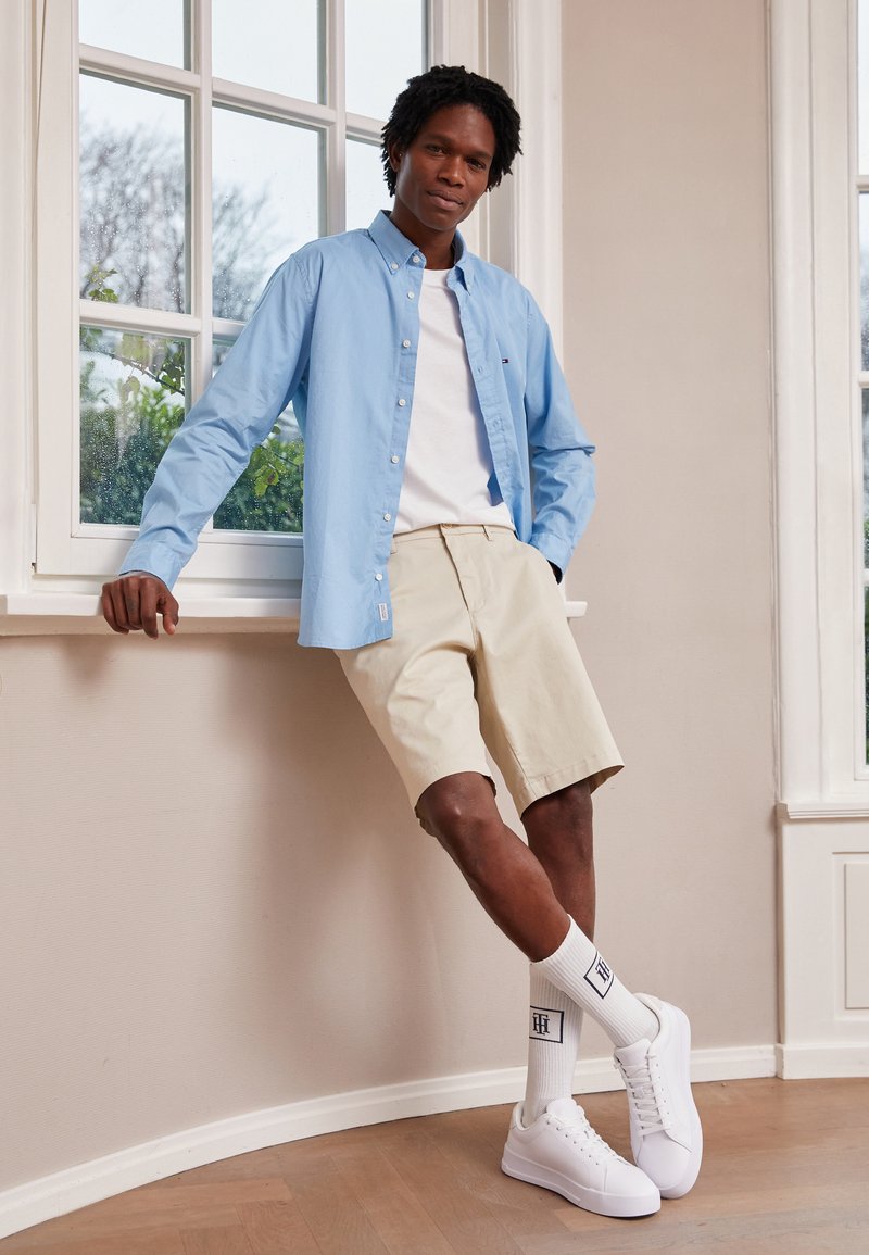 Camicia azzurra a maniche lunghe sopra una maglietta bianca, abbinata a pantaloni corti beige. Sneakers bianche e calze bianche fino al ginocchio con logo.