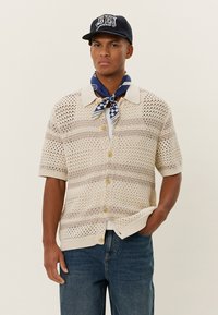 Beige gestricktes Kurzarmhemd mit Lochmuster, ausgestattet mit einem leichten Kragen, Knöpfen und getragen mit einem blauen gemusterten Schal.