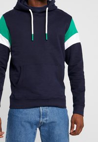 Felpa con cappuccio di colore blu navy con strisce sulle spalle verdi, bianche e blu scuro. Presenta una tasca a marsupio anteriore e cordini bianchi. Materiale morbido con una vestibilità casual.