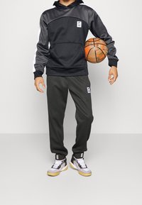 Sweat-shirt à capuche noir avec manches grises, poche kangourou, pantalon de jogging gris et baskets grises et blanches, tenant un ballon de basket orange.