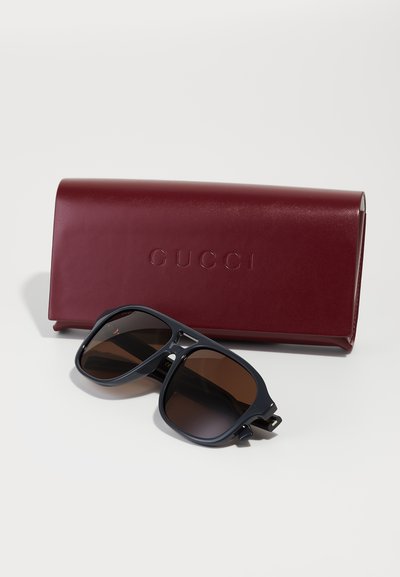 Gafas de sol Gucci con lentes marrón oscuro y montura de plástico azul marino, acompañadas de una funda de cuero burdeos grabada con el nombre de la marca.