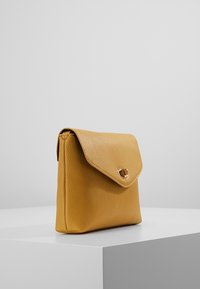 Borsa a tracolla gialla in pelle con finitura testurizzata, chiusura a flap e un accento in metallo dorato sul fronte. Forma rettangolare compatta.
