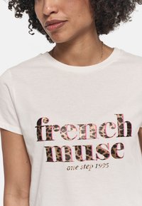 Weißes Baumwoll-T-Shirt mit dem Aufdruck "french muse" im Leopardenmuster, pinken Umrandungen und goldenen Akzenten, kurzen Ärmeln und Rundhalsausschnitt.