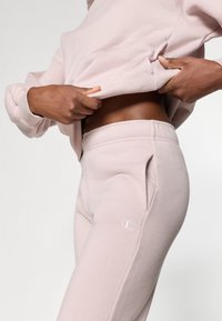 Sweat-shirt en coton rose clair et pantalon de jogging assorti. Présente un logo sur le pantalon, des poignets côtelés et une coupe décontractée. Texture lisse.
