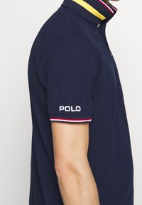 Tmavě modré polo tričko se žlutým límečkem a červeno-bílými pruhovanými detaily, s vyšívaným nápisem "POLO" na levém rukávu.