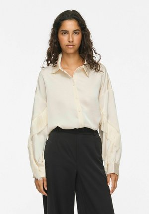 Femme aux cheveux bouclés portant une chemise crème boutonnée avec des détails à franges sur les manches et un pantalon noir taille haute, debout devant un fond uni.