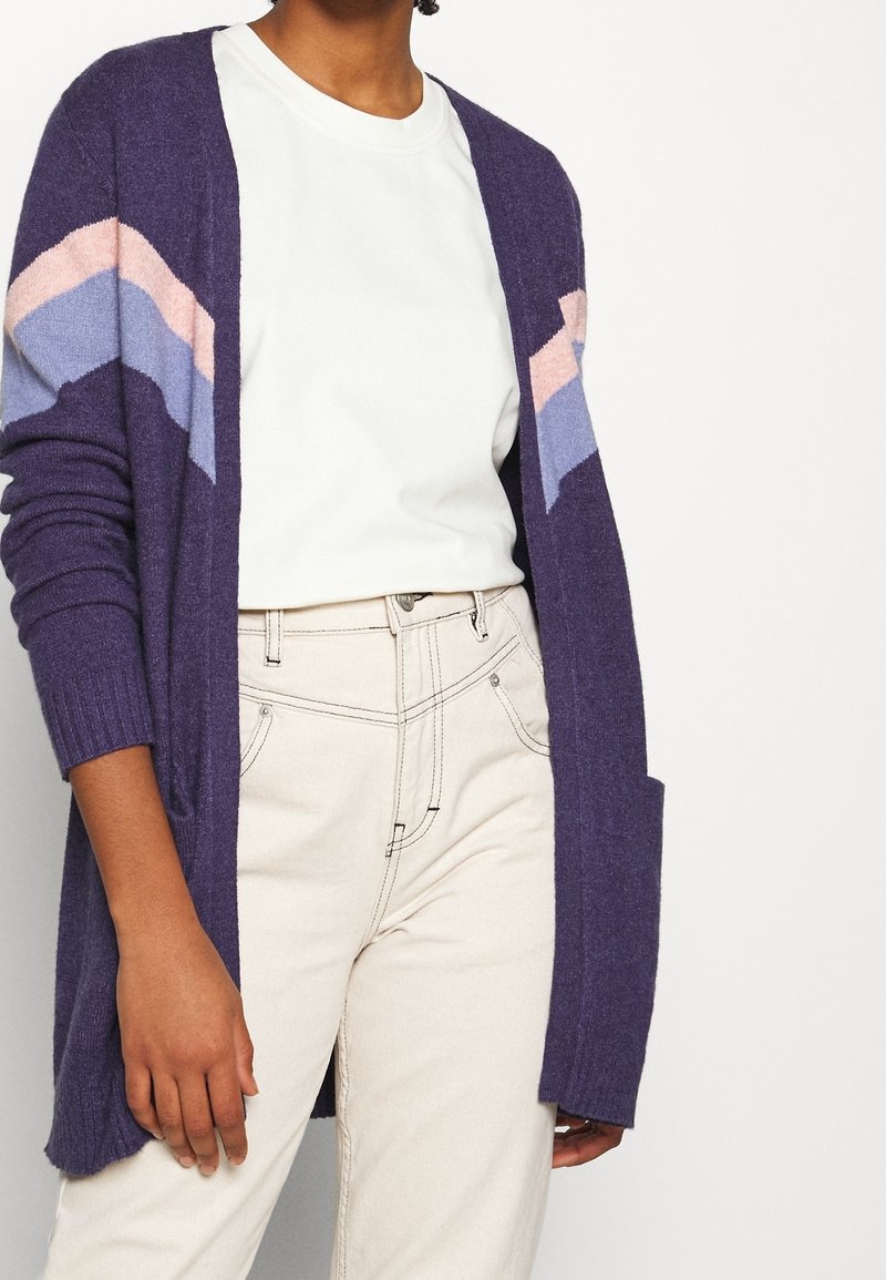 Personne portant un cardigan violet avec des rayures chevrons roses et bleues sur une chemise blanche et un pantalon beige taille haute.