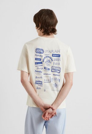Junge Person mit braunen Haaren steht mit dem Rücken zur Kamera, trägt ein weißes T-Shirt mit mehreren blauen "Farah"-Logos und hellblaue Hose.
