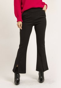 Pantalons évasés noirs avec des fentes latérales et une coupe ajustée, fabriqués à partir d'un tissu extensible. Portés avec un pull en tricot rose vif et des bottines.